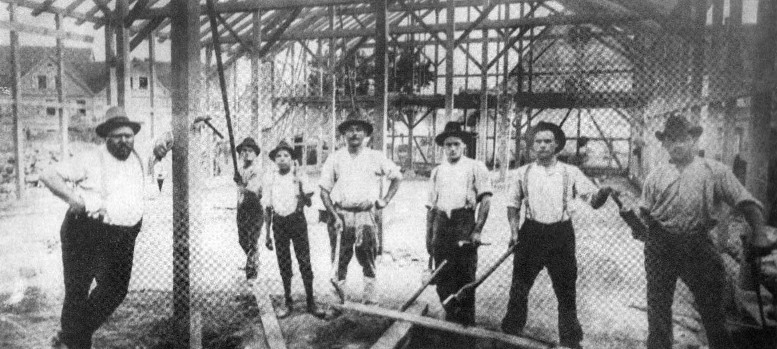 Historisches Foto: acht Zimmermänner, einer ohne Werkzeug in den Händen – vermutlich der Vorarbeiter, stehen in einem Fachwerkhaus-Rohbau aus Holz.