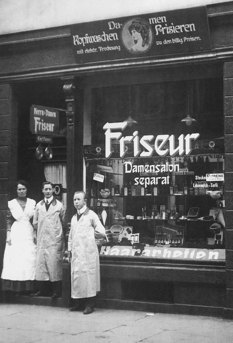 historisches Foto: eine Frau und zwei Männer im Kittel stehen von ihrem Friseur-Laden.